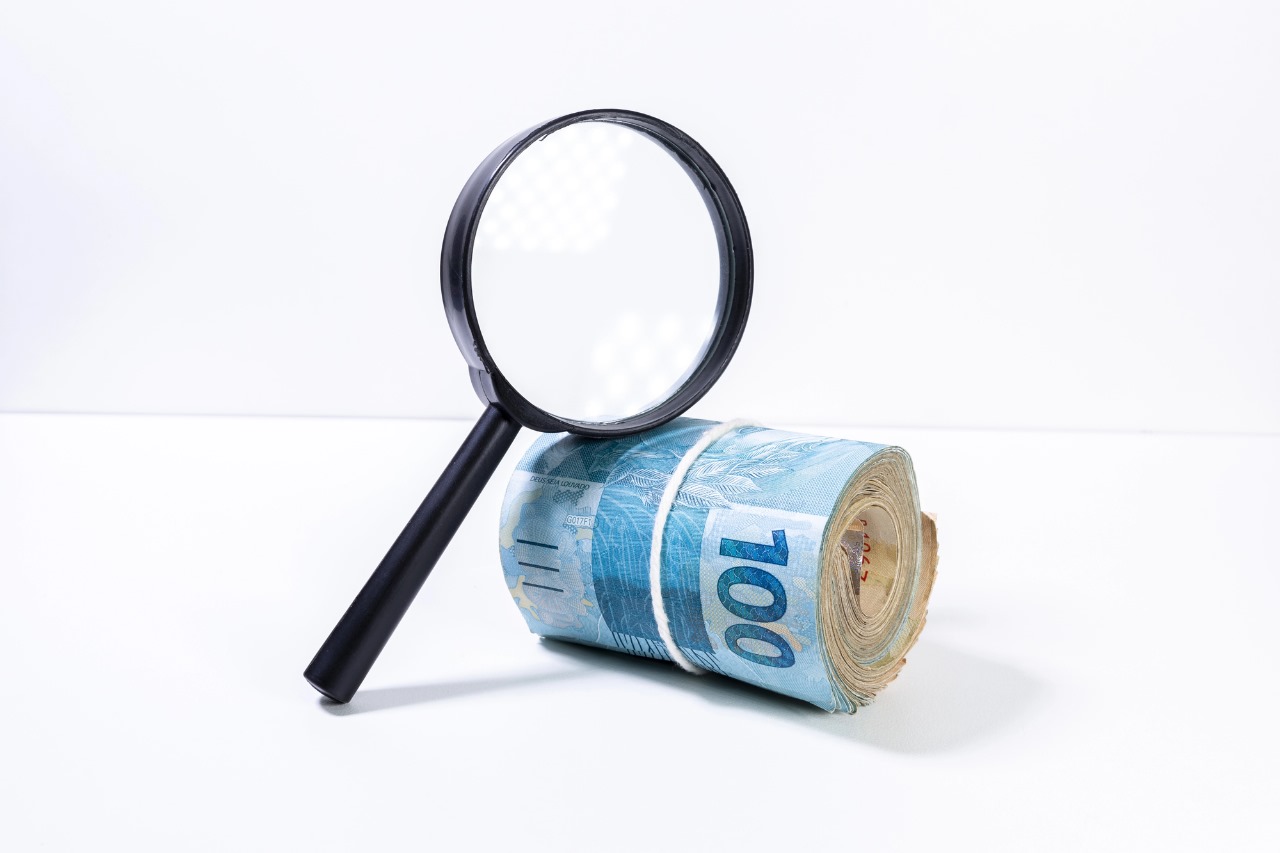 Lupa apoiada em um rolo de dinheiro com notas em real