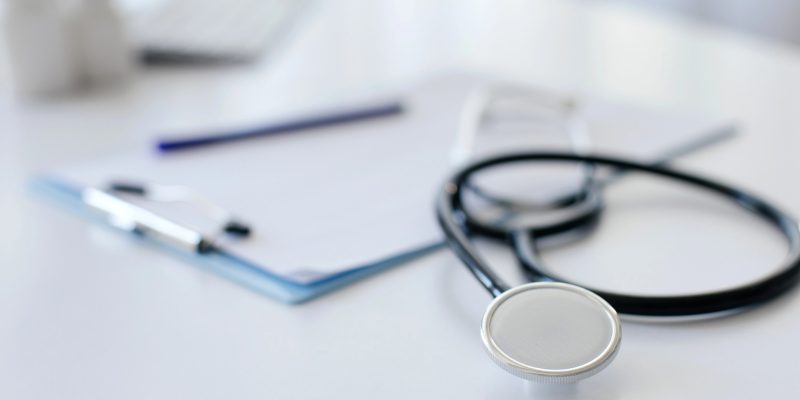 Estetoscópio em primeiro plano sobre uma mesa branca ao lado de uma prancheta com papéis e caneta.
