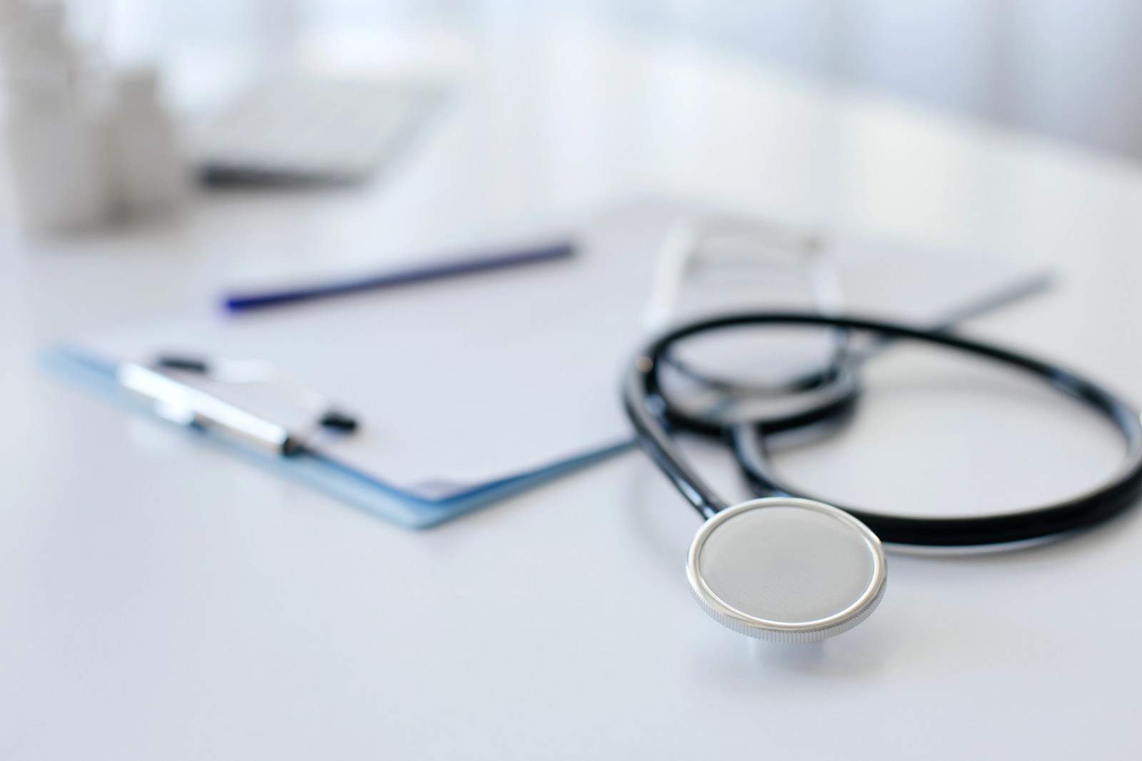 Estetoscópio em primeiro plano sobre uma mesa branca ao lado de uma prancheta com papéis e caneta.