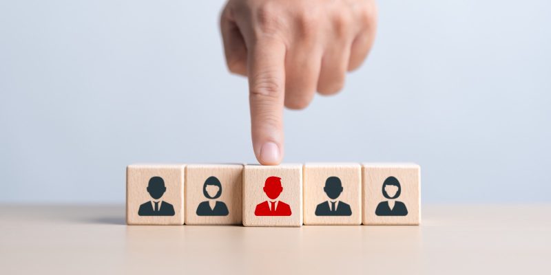 Imagem de uma mão apontando para quadradinhos em madeira, pintados com figuras de homens e mulheres.