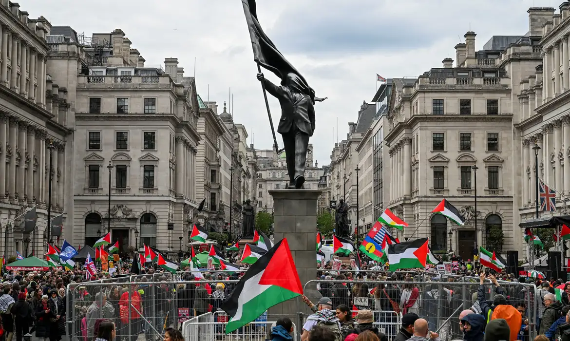 Pessoas seguram bandeiras palestinas ao lado de uma estátua, representando uma figura carregando uma bandeira cobrindo o rosto e descendo de um pedestal, do artista de rua britânico Banksy, durante uma marcha, realizada pela Palestine Solidarity Campaign, para marcar o 78º aniversário da Nakba e contra o comício