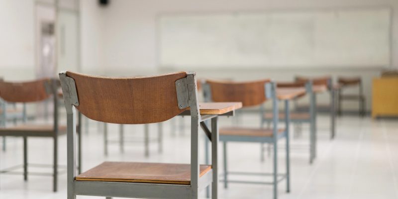 Uma sala de aula vazia com foco em uma cadeira escolar de madeira e metal em primeiro plano, com outras fileiras de carteiras e um quadro branco ao fundo de forma desfocada.