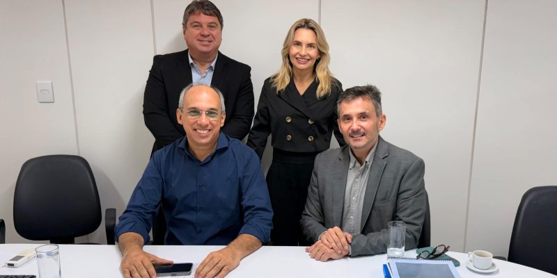 Quatro pessoas, sendo três homens e uma mulher, sorriem para a foto em um ambiente de escritório, com dois homens sentados à mesa e o outro casal em pé logo atrás.