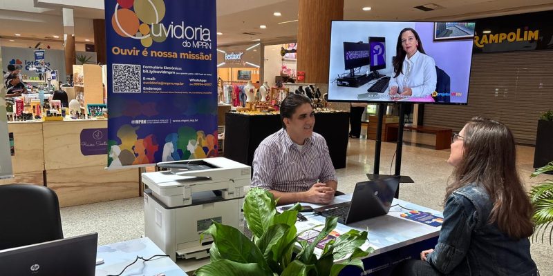 Homem e mulher conversam sentados em um estande de atendimento da Ouvidoria do MPRN, estruturado com computadores, impressora e um painel informativo, no corredor de um shopping.