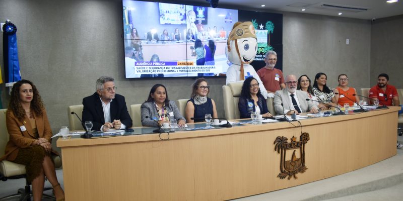 audiência pública em um auditório, mostrando uma mesa com diversas autoridades sentadas e um boneco mascote em pé ao fundo, posicionados em frente a um grande painel que exibe a transmissão do evento.
