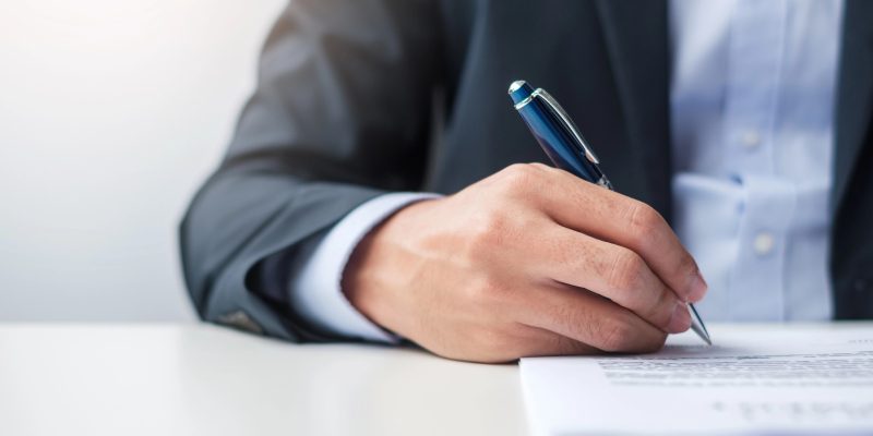 Homem em traje formal segura uma caneta azul e assina um documento sobre uma mesa branca.