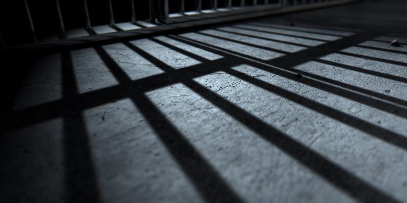 Sombra de grades de prisão projetada sobre o chão de concreto de uma cela escura.