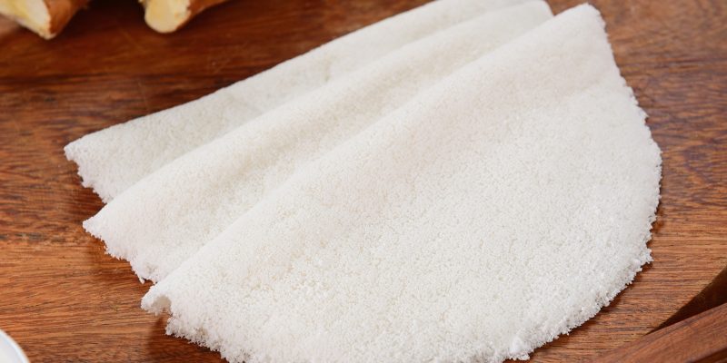 Uma tapioca branca, dobrada ao meio em formato de meia-lua, está disposta sobre uma tábua de madeira escura com pedaços de mandioca ao fundo.