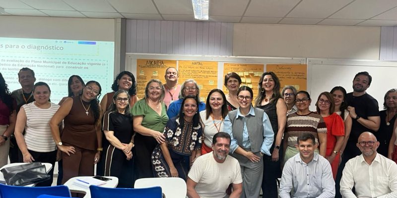 Grupo de cerca de vinte pessoas posa sorridente para foto em uma sala de aula com carteiras escolares azuis em primeiro plano e uma tela de projeção ao fundo.