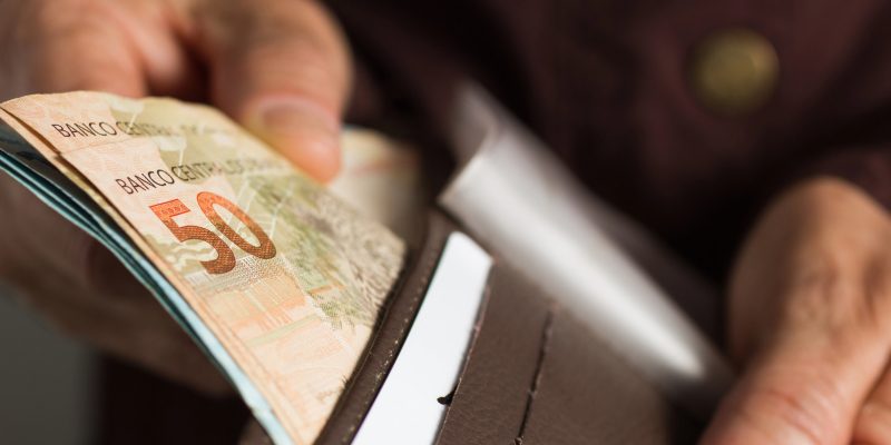 close mostra as mãos de uma pessoa idosa retirando uma nota de 50 reais de dentro de uma carteira marrom aberta