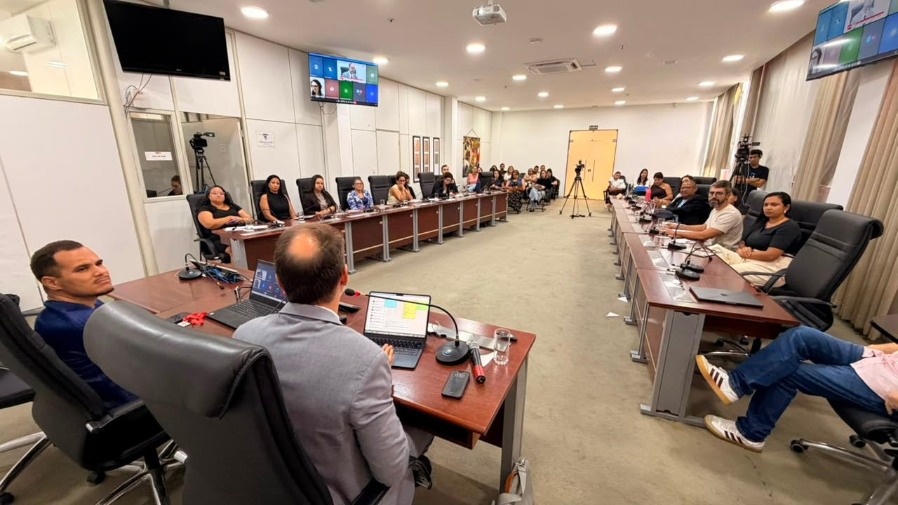 Pessoas reunidas em uma sala de conferências, dispostas ao redor de grandes mesas organizadas em formato de U, acompanhando uma reunião com transmissão ao vivo.