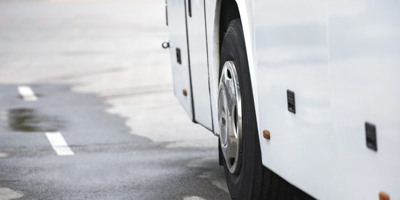 lateral inferior de um ônibus branco estacionado sobre o asfalto molhado, destacando um de seus pneus com calota cromada
