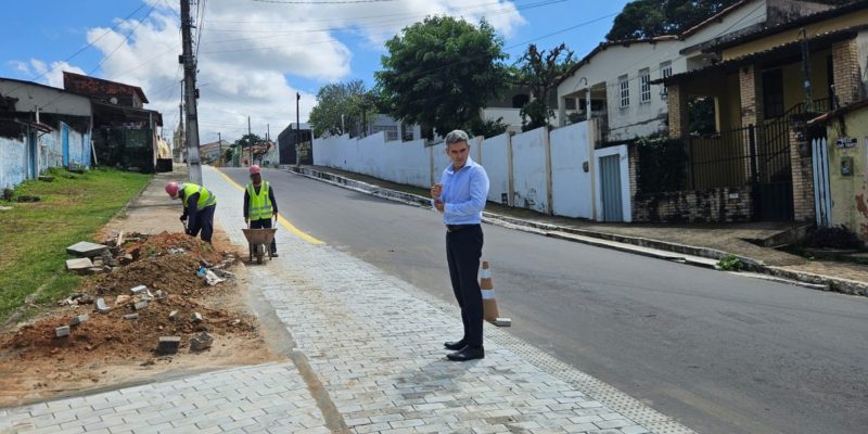 Homem de camisa social clara e calça escura observa dois operários trabalhando na pavimentação de uma calçada em uma rua residencial ladeada por casas e árvores sob céu ensolarado.