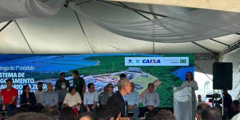 promotora discursa em um púlpito durante um evento oficial sob uma tenda, acompanhada por autoridades no palco e com uma grande plateia assistindo.
