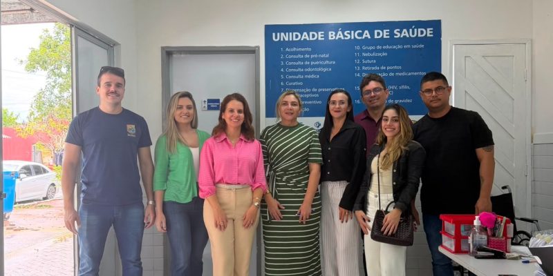 Um grupo de oito pessoas sorri em pé, lado a lado, dentro de uma Unidade Básica de Saúde, posando para uma foto em frente a uma placa azul com a lista de serviços oferecidos pelo local.