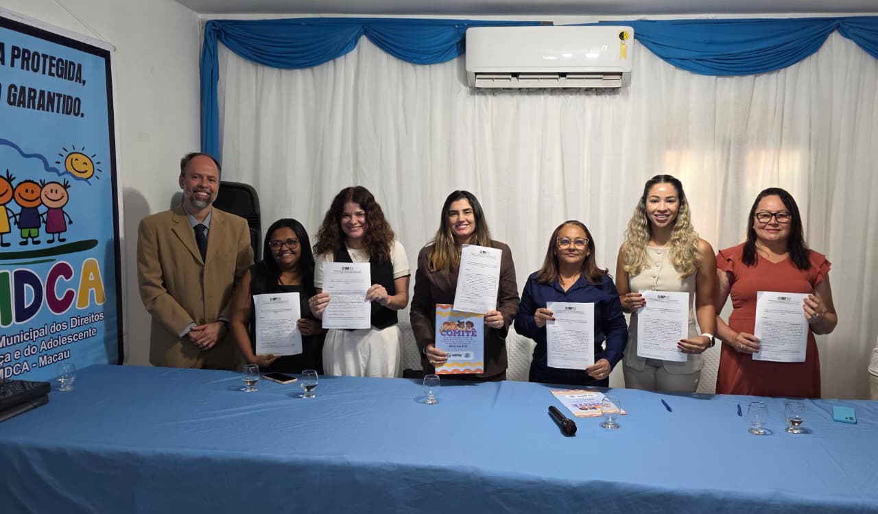 Sete pessoas estão lado a lado atrás de uma mesa azul, sorrindo e segurando documentos impressos em um evento oficial do CMDCA de Macau.
