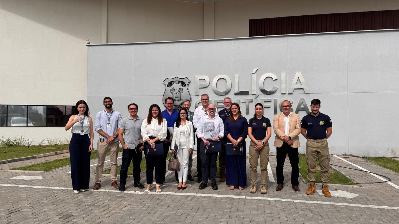 grupo de treze pessoas sorri e posa para foto em frente ao prédio da Polícia Científica.