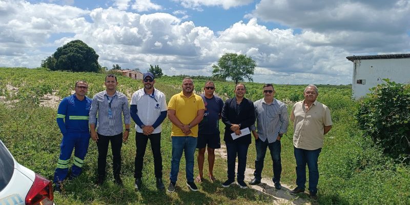Grupo de oito homens em pé lado a lado em uma área rural ao ar livre, com vegetação e céu ensolarado ao fundo.