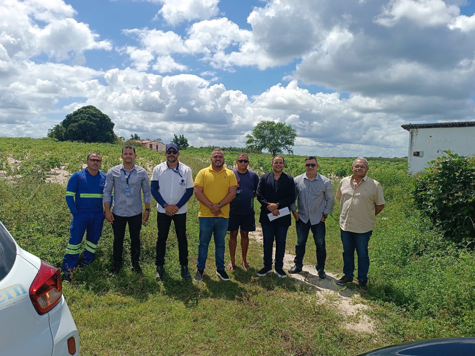 Grupo de oito homens em pé lado a lado em uma área rural ao ar livre, com vegetação e céu ensolarado ao fundo.