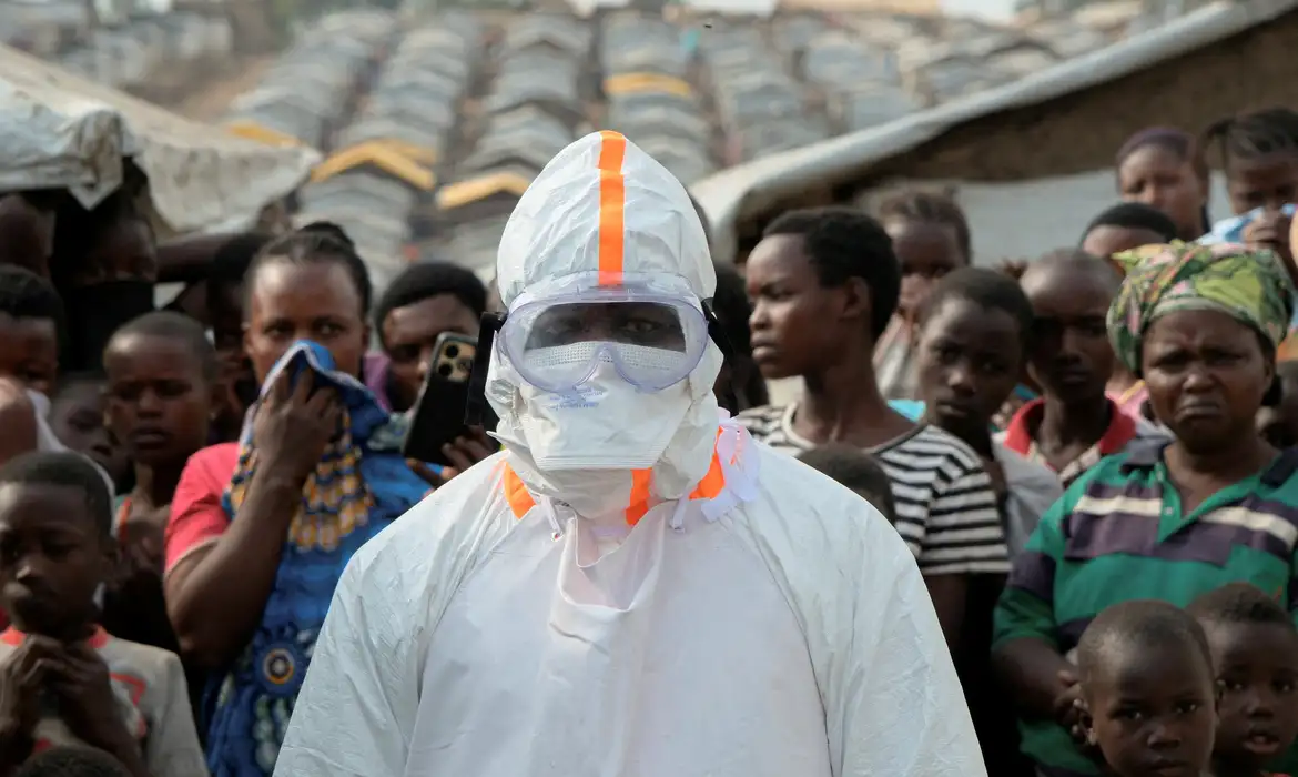 Profissional de saúde usando equipamento de proteção individual ao lado de pessoas deslocadas que aguardam enterro de vítimas com suspeita de Ebola no campo de deslocados de Kigonze, em Bunia, no leste da República Democrática do Congo
18 de junho de 2026 REUTERS/Gradel Muyisa Mumbere