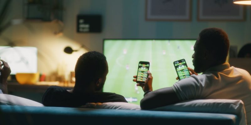 13/07/2026 - Bets - African American man using smartphone and clinking bottles of beer with friends while chilling on sofa and watching soccer game on TV. Foto: Framestock/ Adobe Stock