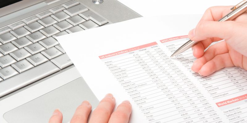 imagem mostra as maos de uma pessoa conferindo uma lista em papel com um computador ao fundo