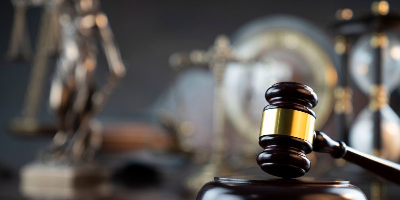 martelo de juiz em madeira escura e detalhes dourados se destaca em primeiro plano, tendo ao fundo, de forma desfocada, símbolos da justiça como a estátua da Temis e uma ampulheta