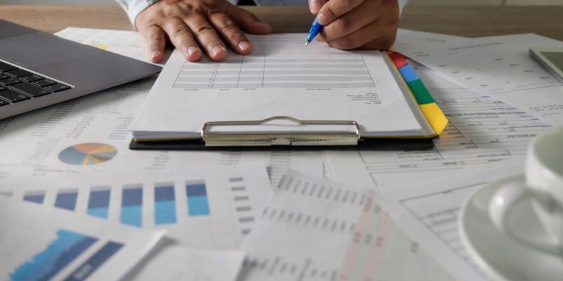Pessoa escrevendo em documentos sobre uma mesa de escritório repleta de relatórios, gráficos financeiros e um notebook.