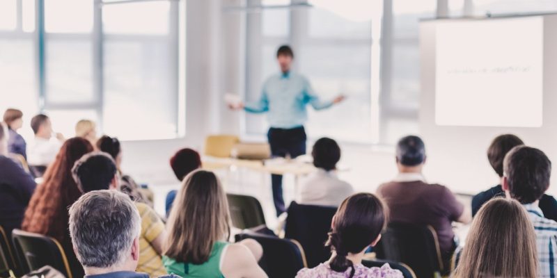 Fotografia em plano geral de uma plateia de costas assistindo a uma palestra ministrada por um homem em uma sala iluminada.