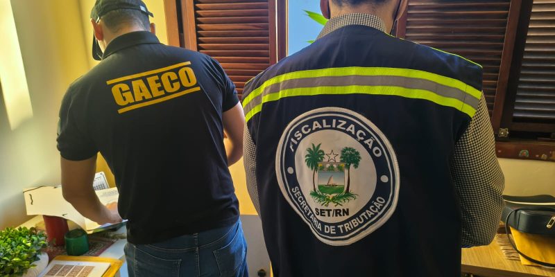 Dois agentes de fiscalização de costas, um usando uniforme do GAECO e o outro do SET/RN, examinam documentos em uma sala.