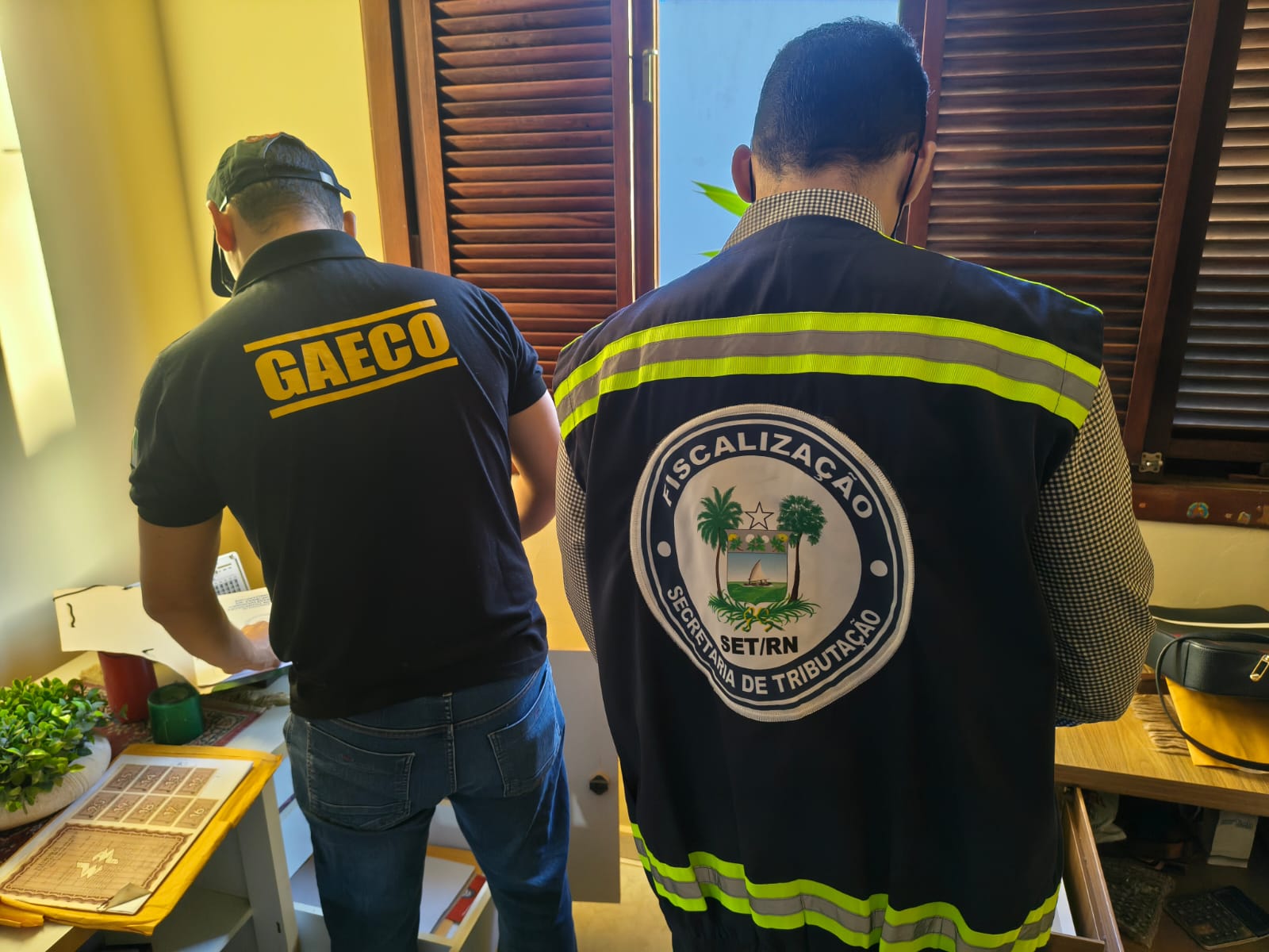 Dois agentes de fiscalização de costas, um usando uniforme do GAECO e o outro do SET/RN, examinam documentos em uma sala.