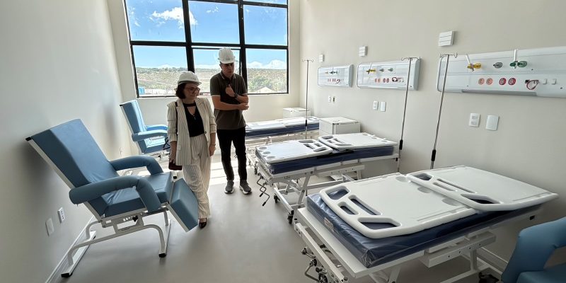 Duas pessoas usando capacetes brancos de proteção inspecionam um quarto hospitalar novo e iluminado, equipado com poltronas azuis e várias camas hospitalares dispostas em fila.