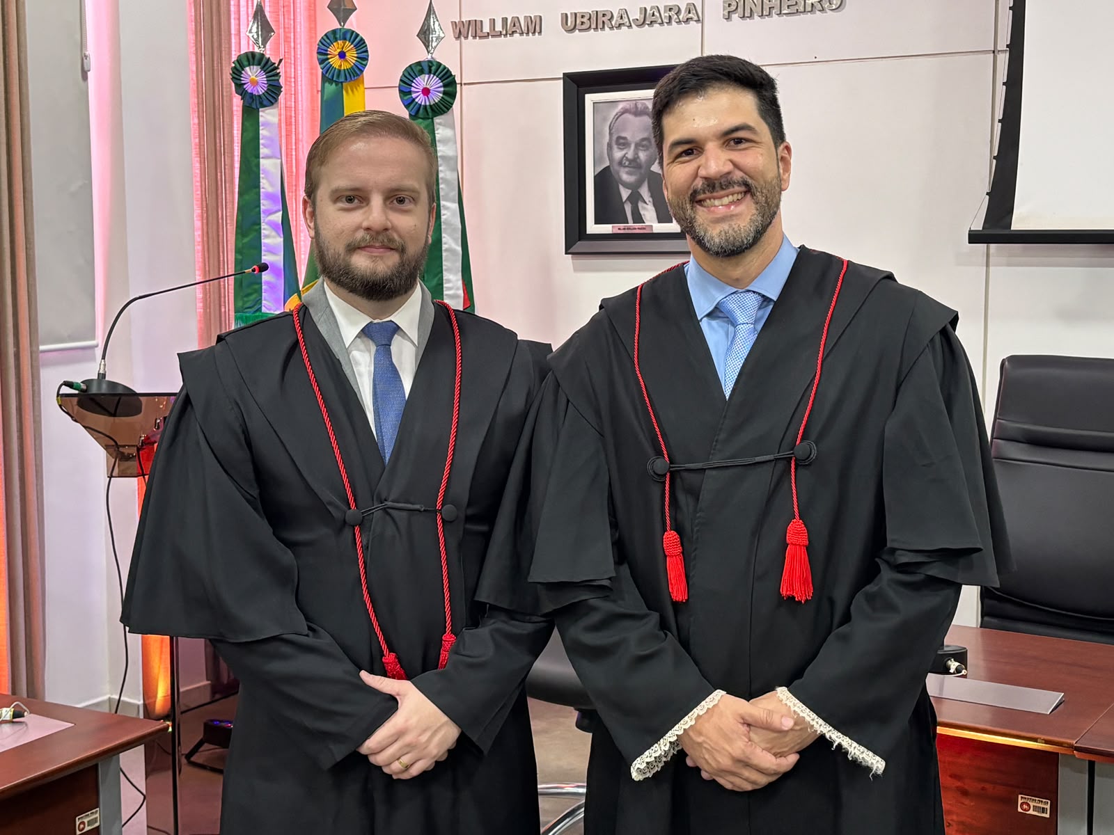 Dois homens vestidos com togas pretas estão em pé e posam para uma foto em um ambiente de escritório com bandeiras ao fundo.